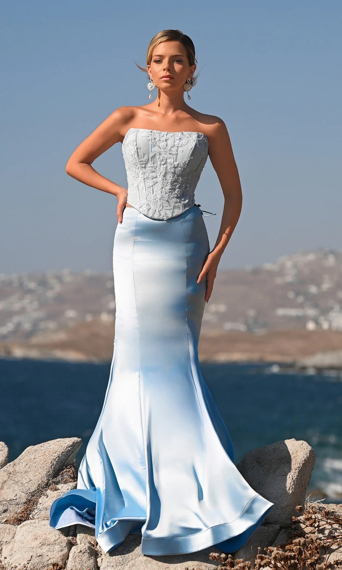 A woman stands on rocks by the water, wearing the Portia and Scarlett PS26092 long formal dress in blue. The strapless gown features a fitted bodice and flared skirt, with hazy hills and a blue sky creating a dreamy backdrop.